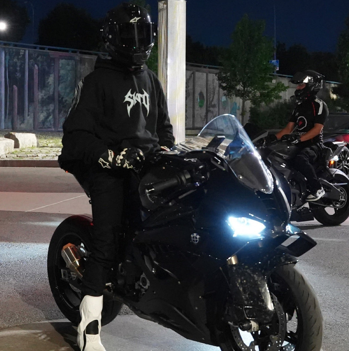 Helmeted rider in a black hoodie with white chest graphic on a parked black motorcycle at night.