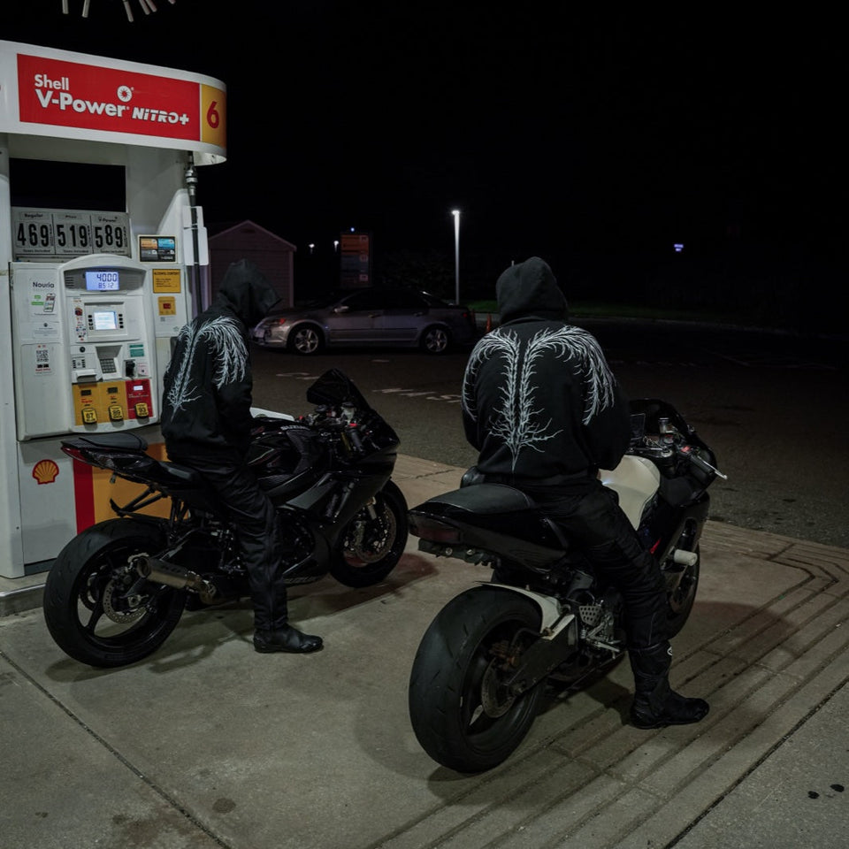 Two helmeted wearers in AUXESIS hoodies beside parked motorcycles.