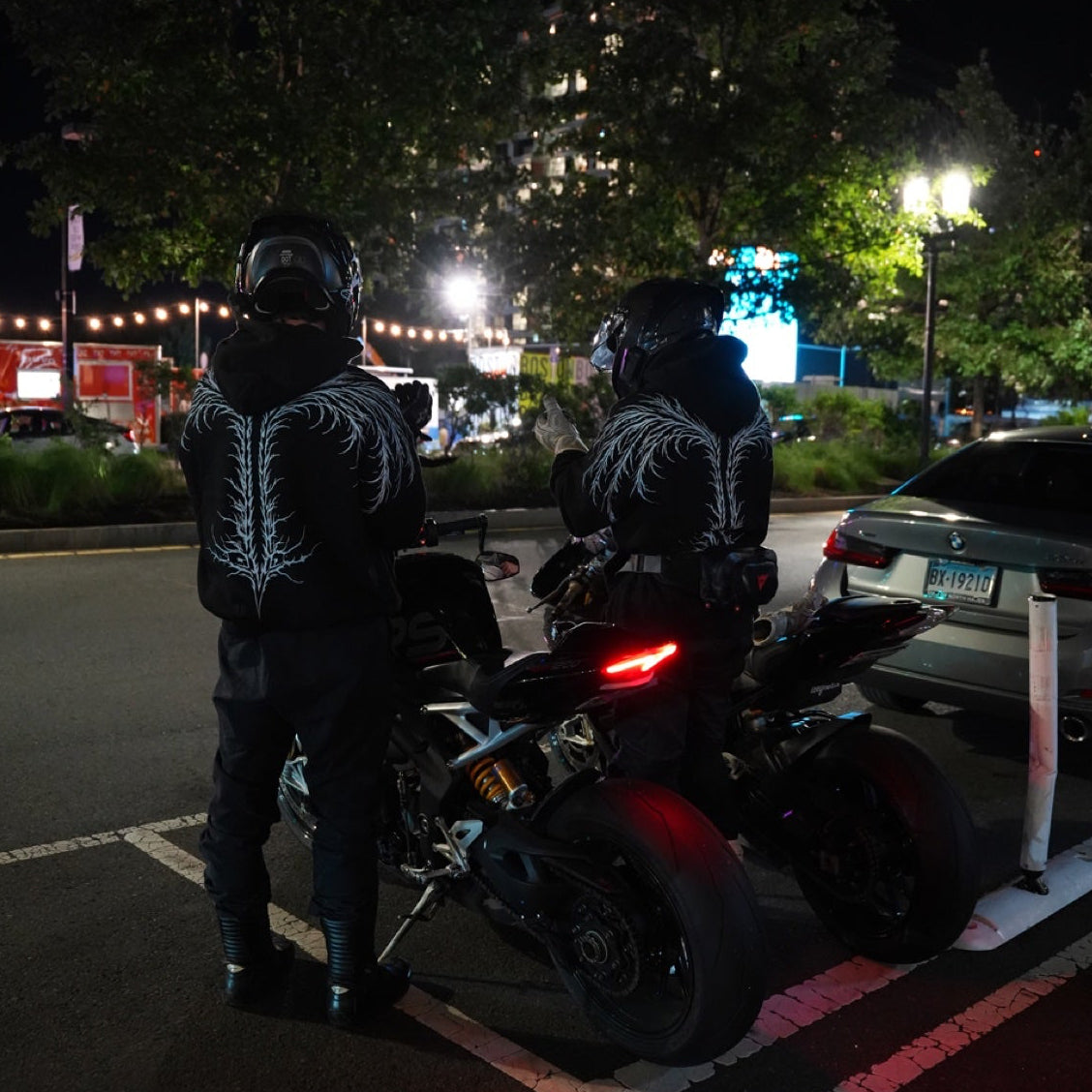 Two helmeted riders wearing black hoodies with white back artwork beside motorcycles on a city street.
