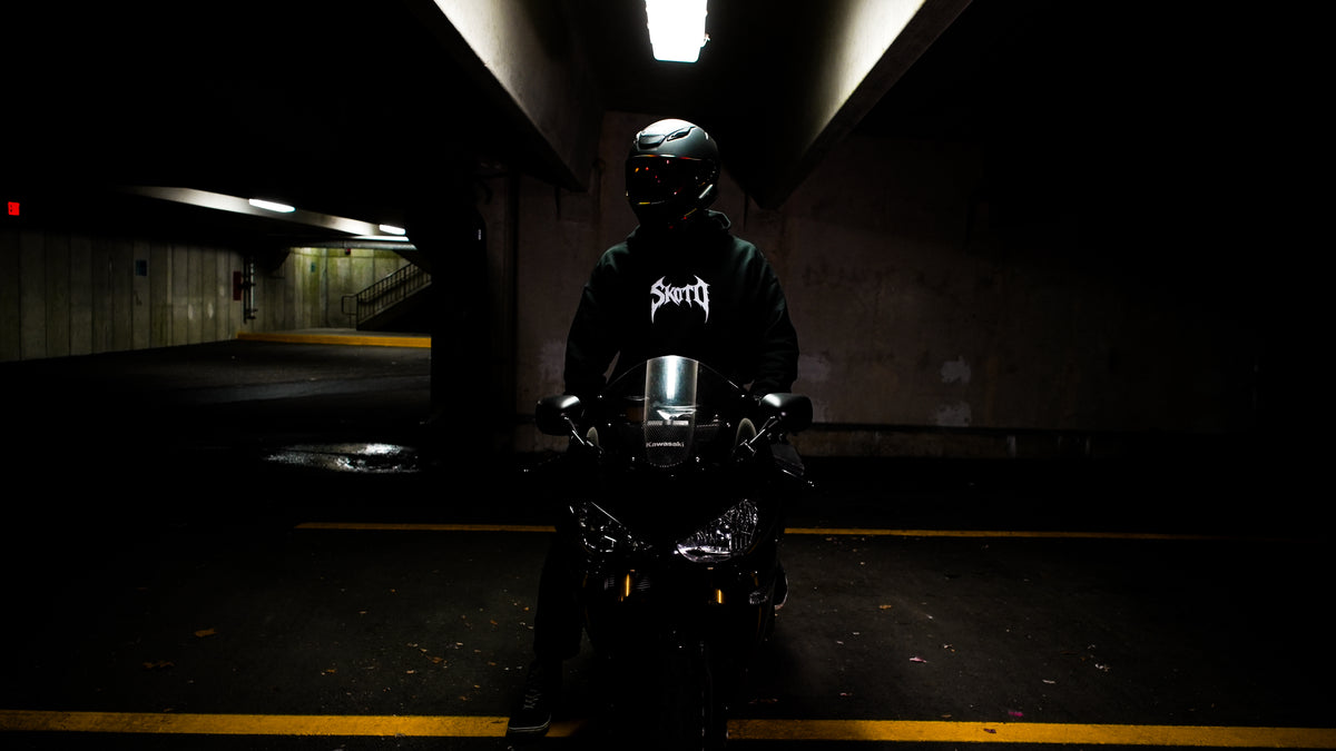 Helmeted rider wearing a black SKOTO hoodie sits on a motorcycle inside a dim concrete garage.