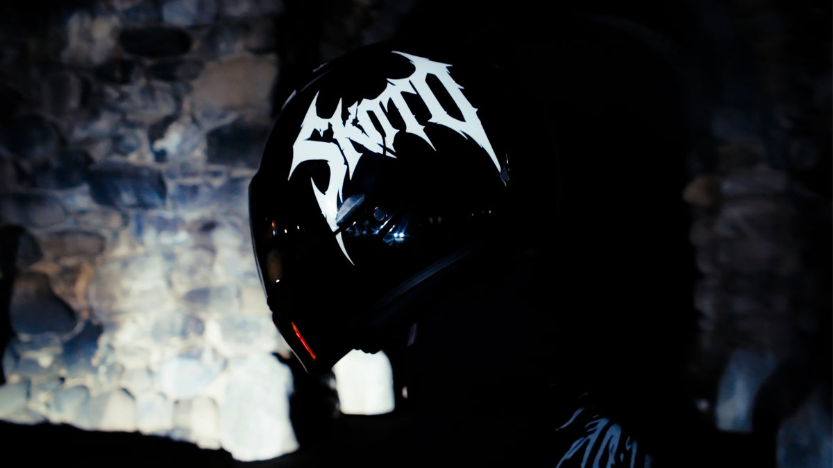 Close view of a black motorcycle helmet with a white SKOTO decal against a textured wall.