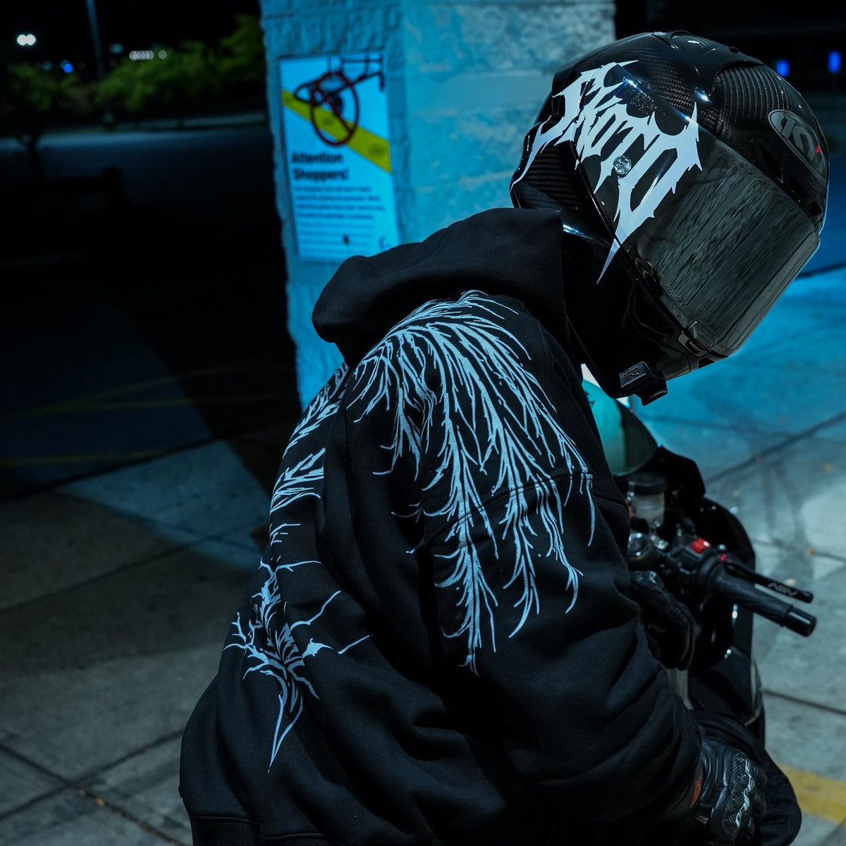Helmeted rider in a black hoodie with white shoulder and back artwork beside a motorcycle under blue night lighting.