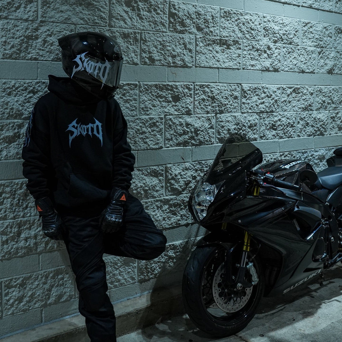 Helmeted rider in a black hoodie standing beside a black sportbike against a cool-lit concrete wall.