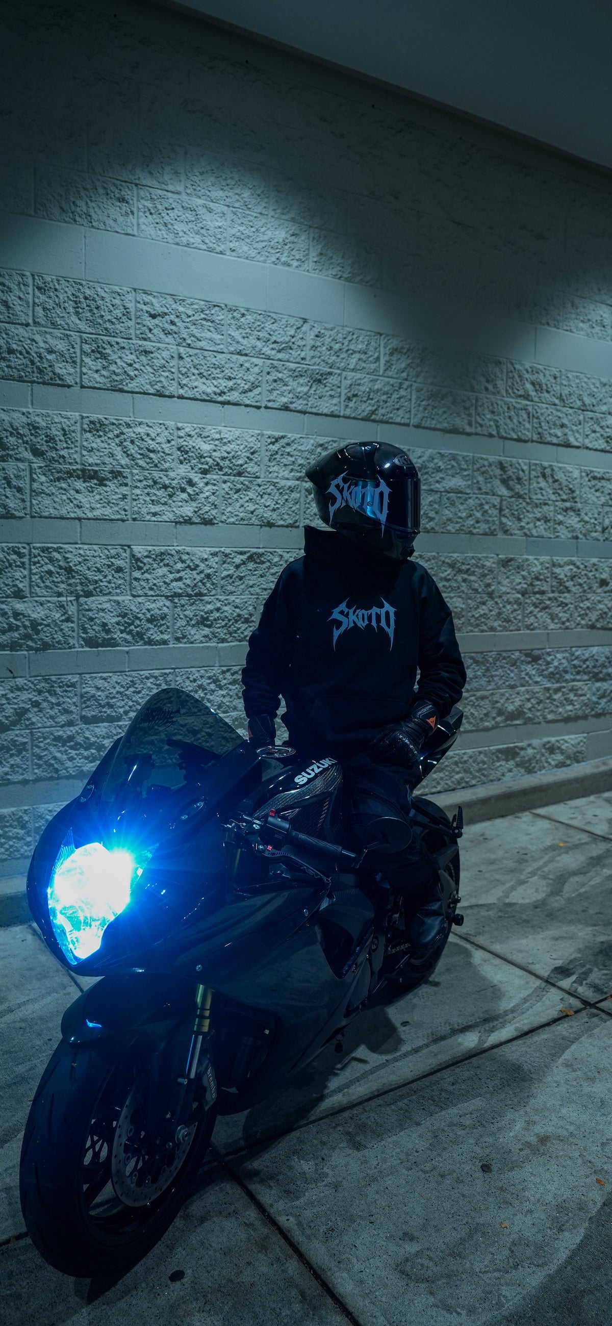 Rider wearing a black hoodie on a blue-lit motorcycle beside a textured wall at night.