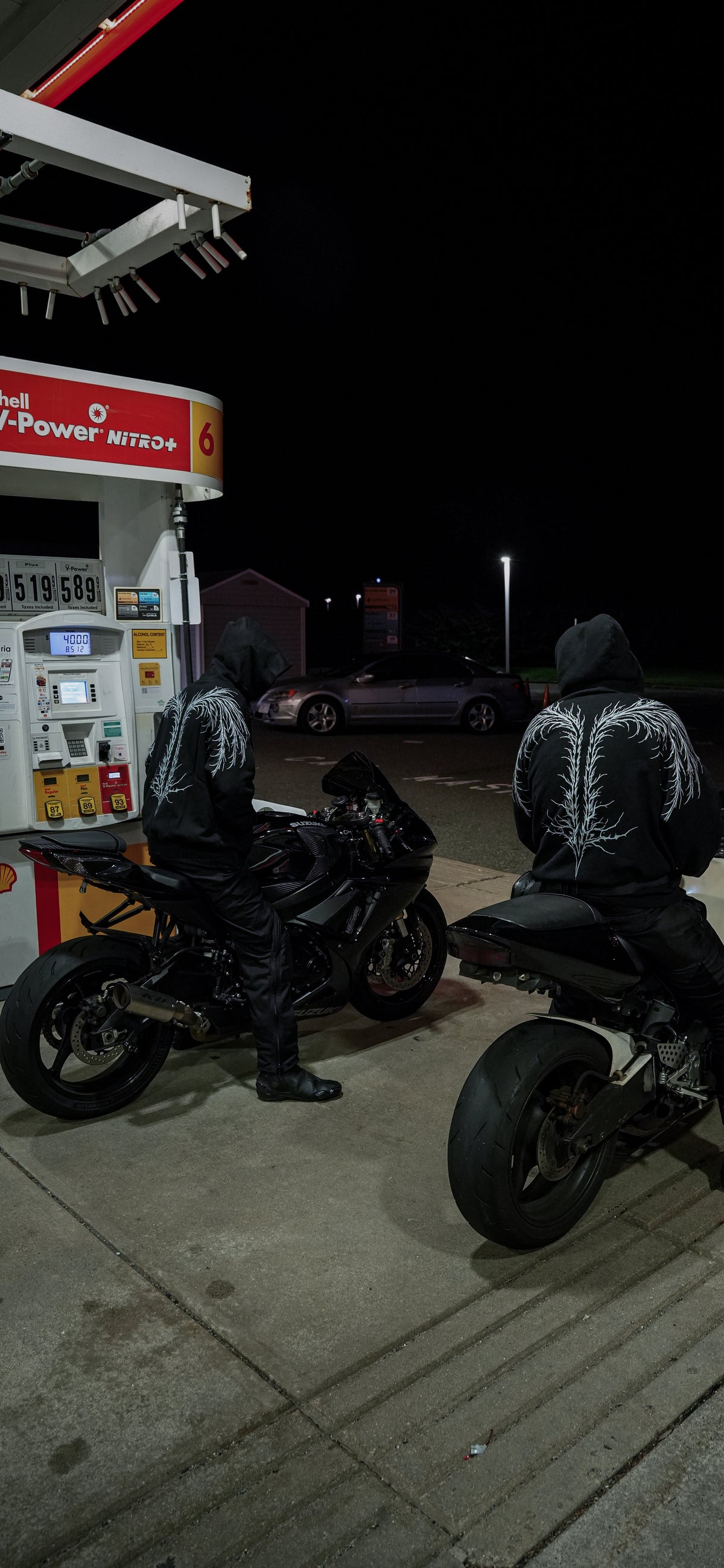 Two helmeted riders in black hoodies with white back artwork beside motorcycles at a gas station after dark.