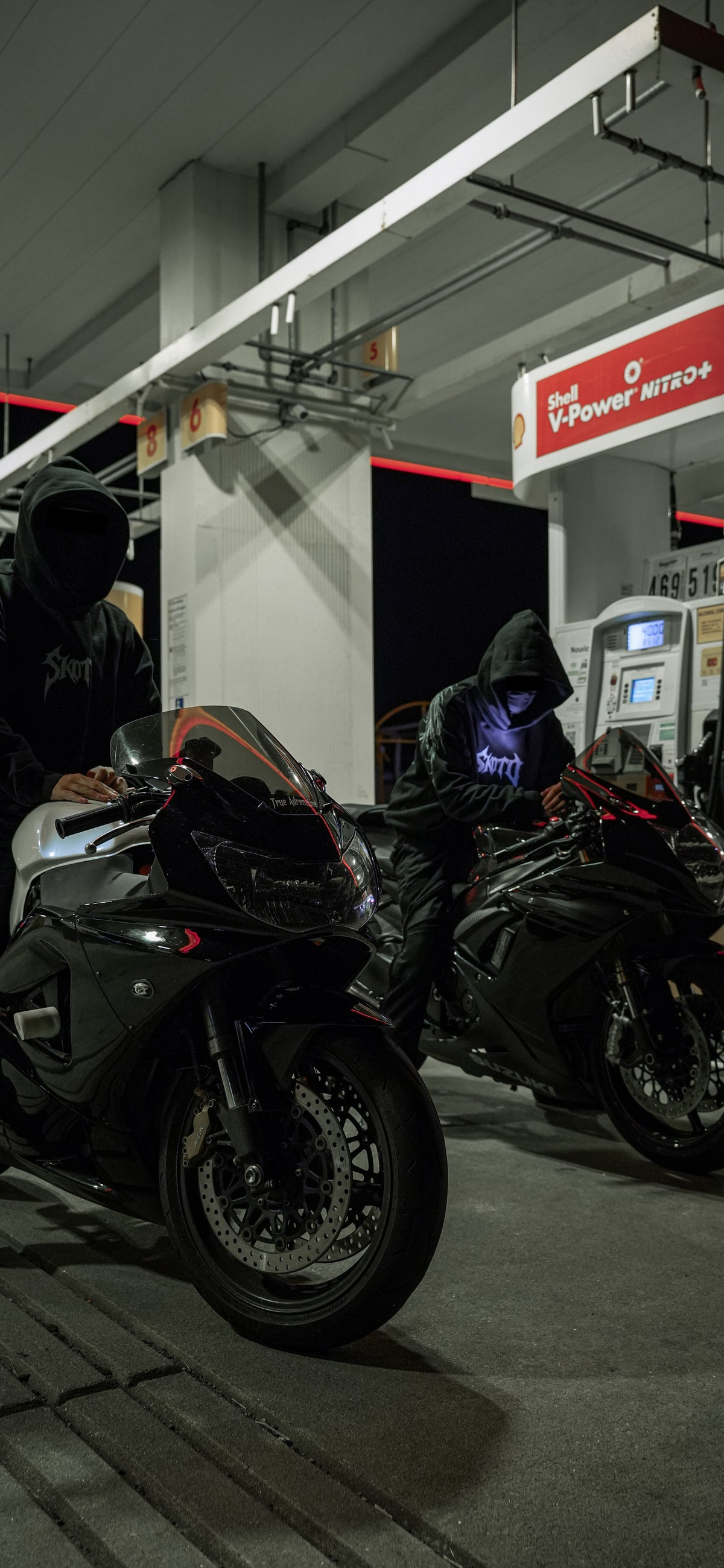 Two riders in black hoodies with sportbikes beneath a gas-station canopy at night.