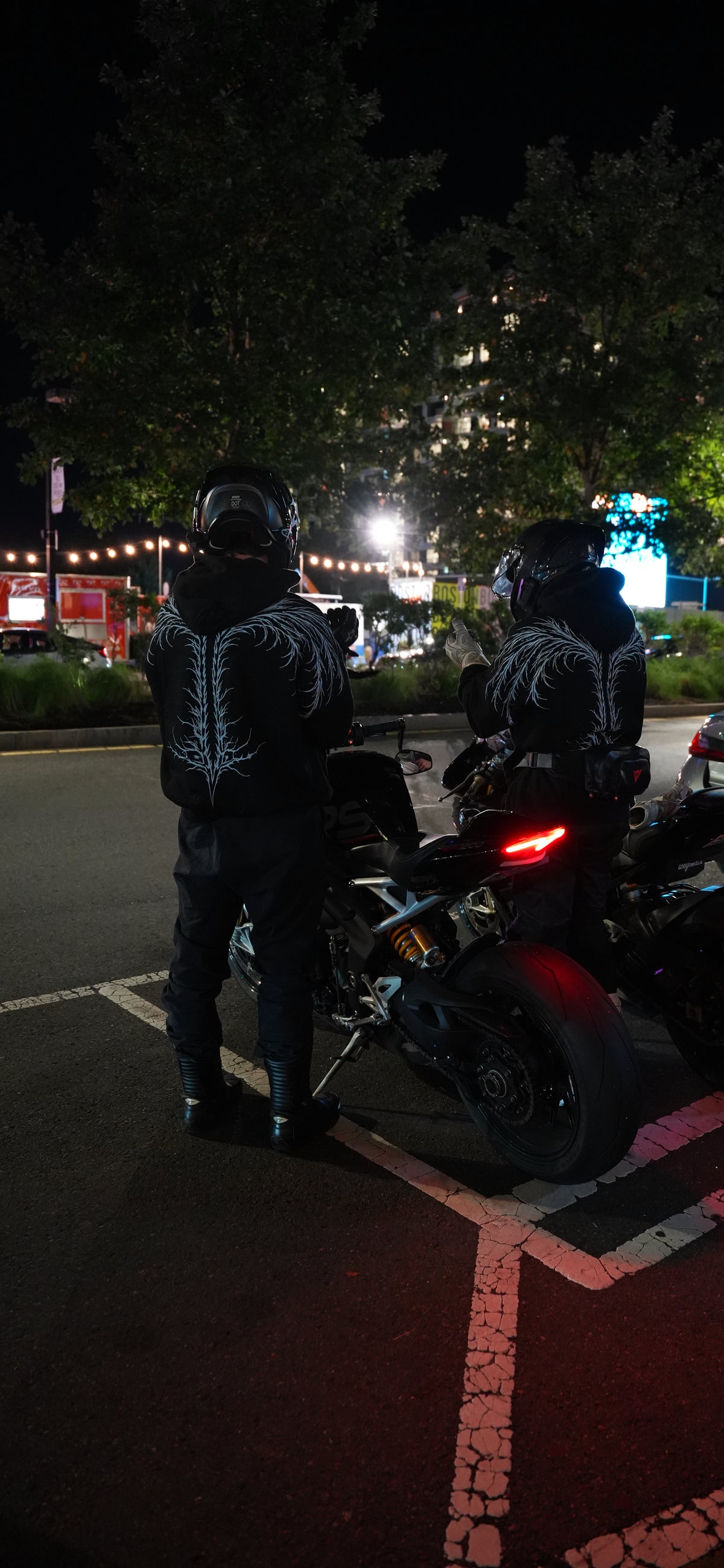 Two helmeted riders in black hoodies with white back artwork stand beside motorcycles on a city street at night.