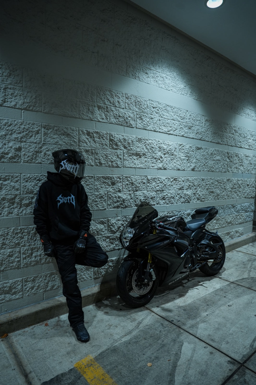 Standing helmeted rider in black hoodie beside a parked sportbike and concrete wall.