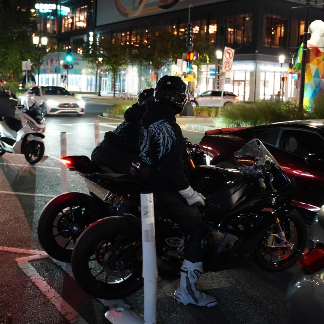 Helmeted riders and parked motorcycles beside cars and illuminated city buildings at night.